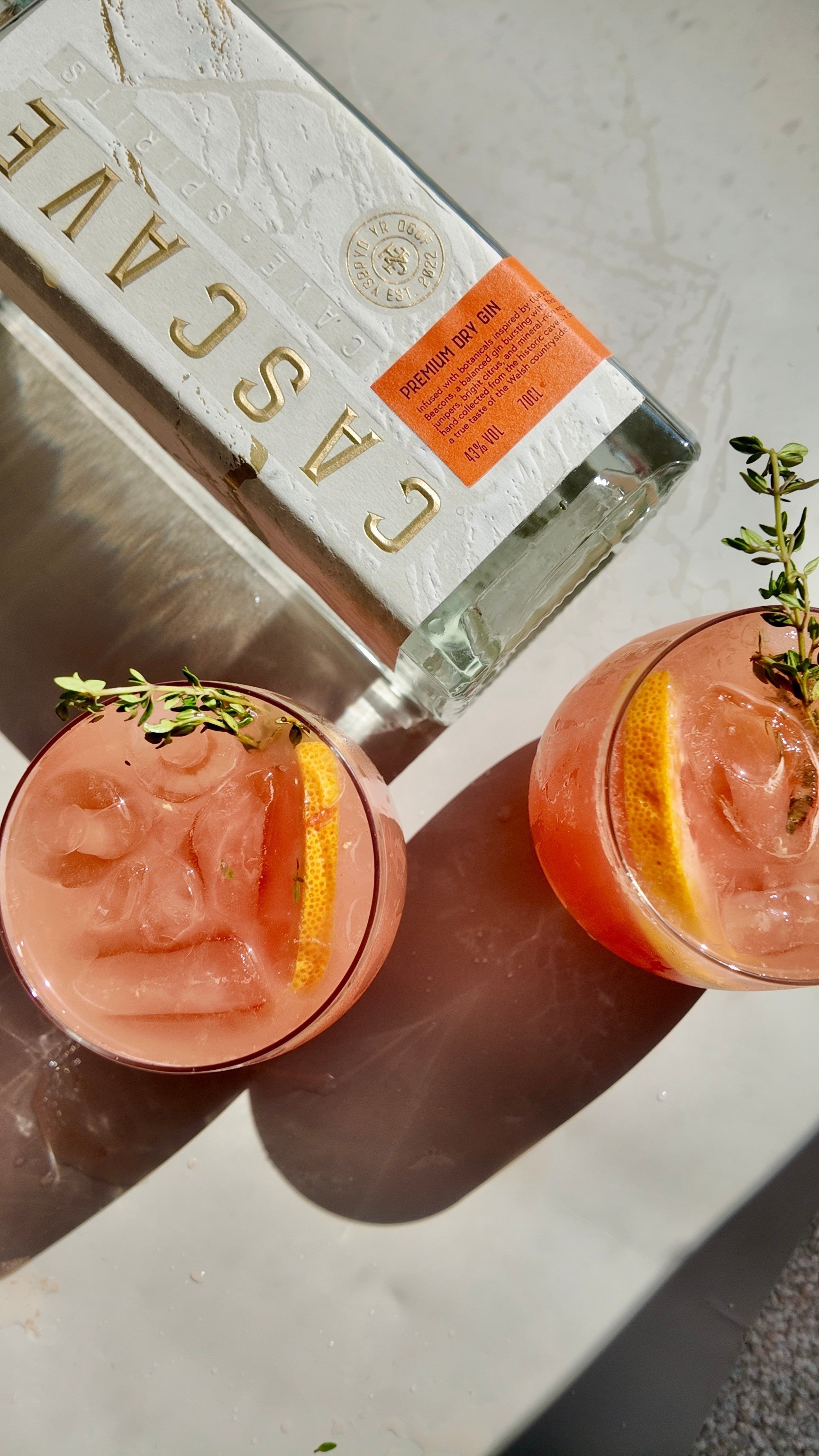 Two pink cocktails with lemon slices and thyme garnish on a marble surface, with a bottle of Cascave Premium Dry Gin in the background.