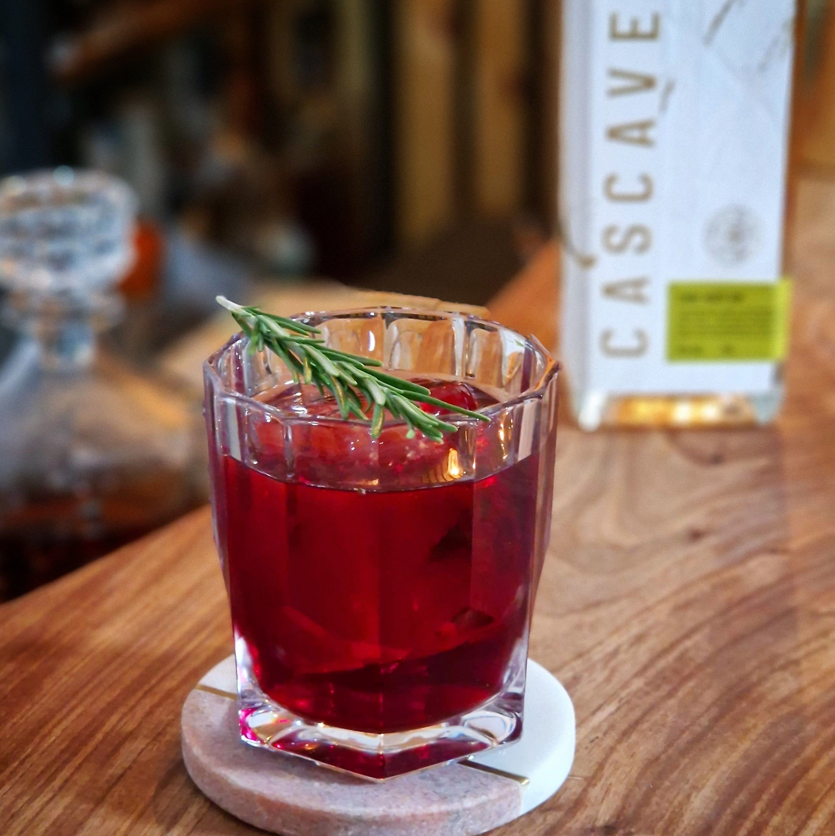 Red cocktail with a sprig of rosemary in a glass on a wooden coaster, with a bottle labeled 'Cascave' in the background.
